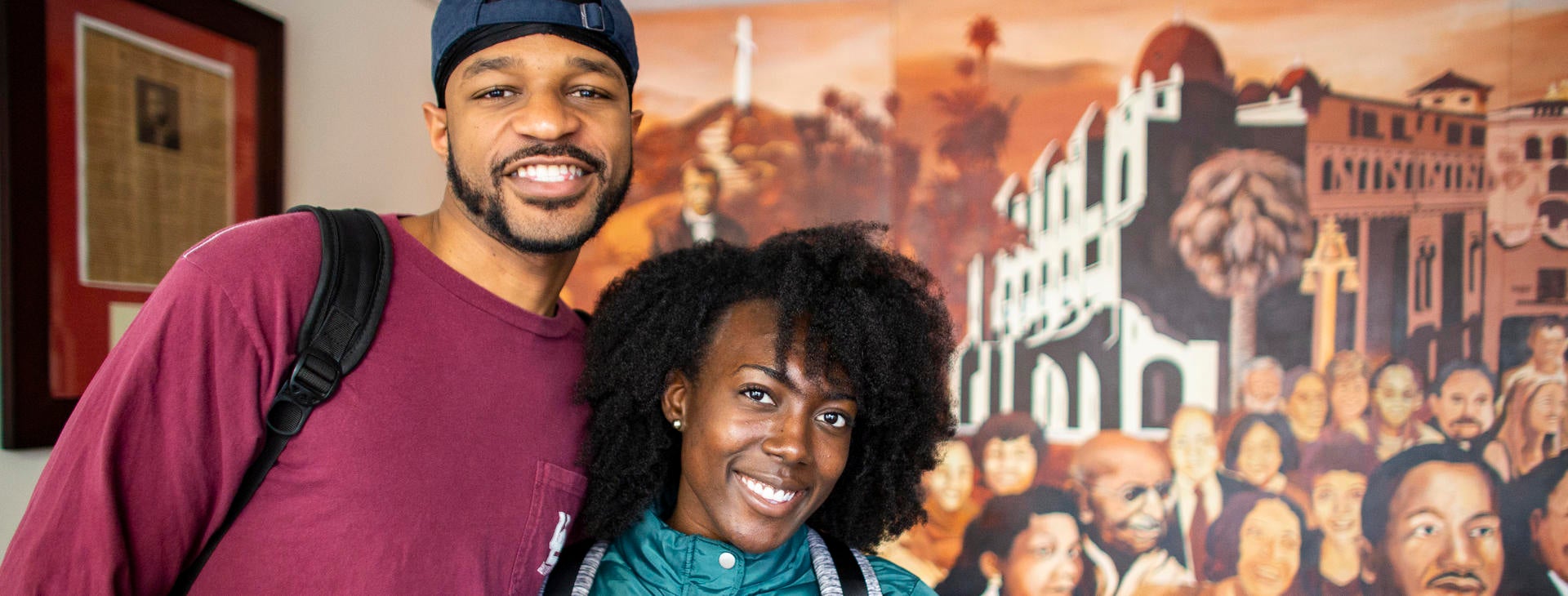 Two students smiling in front of a mural of black leaders