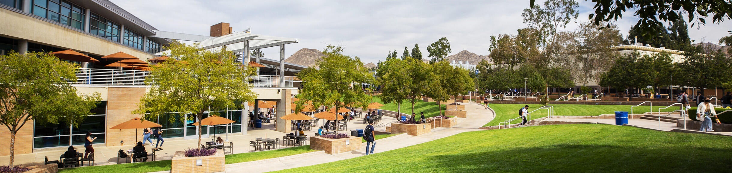 A partly cloudy day on the UC Riverside campus with the Highlander Union Building (HUB) in the background and students sitting at umbrella-covered tables in the HUB plaza.