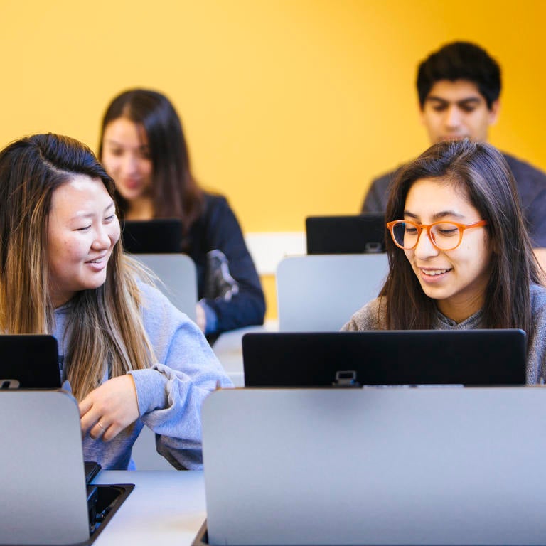 Two students with computers smiling against yellow background.