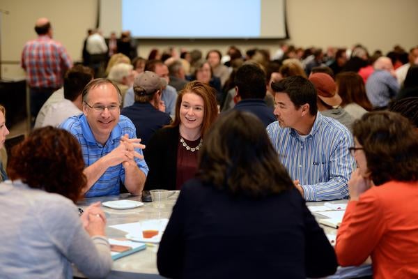 Image of faculty and staff members talking together at a round table in a room full of poeple sitting together at round tables